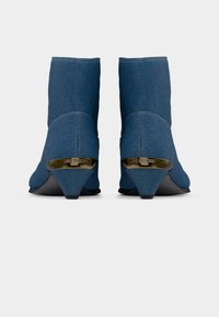 Ζευγάρι μπλε τζιν μποτάκια αστραγάλου με χαμηλά τακούνια τύπου block, χρυσή μεταλλική λεπτομέρεια με εγχάραξη του ονόματος της μάρκας στο πίσω μέρος του τακουνιού.