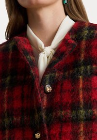 Sarkans un melns ģeometrisks mēteli, izgatavots no teksturētas vilnas, ar zelta krāsas pogām un krēmkrāsas kakla šalli.