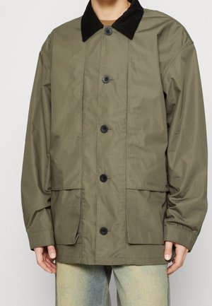 Mężczyzna ubrany w oliwkowozieloną zapinaną na guziki kurtkę z czarnym kołnierzem i dużymi przednimi kieszeniami, stojący na jednolitym jasnym tle.