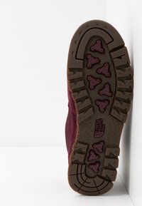 Bordeauxrode zool van een wandelschoen, voorzien van een bruine rubberen buitenzool met driehoekige noppen voor grip. Gestructureerde afwerking en "The North Face" logo aanwezig.