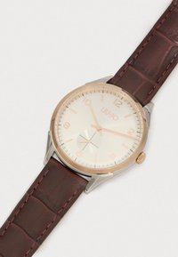 Στρογγυλό ρολόι με λευκό καντράν και ροζ χρυσούς δείκτες. Ο καφέ δερμάτινος ιμάντας διαθέτει υφή κροκοδείλου, ατσάλινη θήκη σε ασημί απόχρωση και κορώνα.