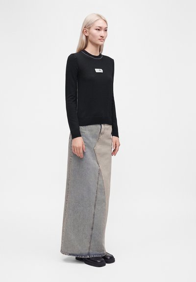 Femme aux longs cheveux blonds portant un haut noir à manches longues et une jupe longue bicolore grise et beige avec des chaussures noires.