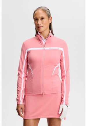 Eine Frau mittleren Alters trägt eine rosa Jacke mit Reißverschluss und weißen Streifen sowie einen passenden rosa Rock. Sie hält einen weißen Golfhandschuh in der Hand.