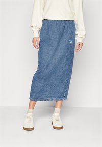 Jupe maxi en denim bleu moyen, dotée d'une taille élastique et d'une patch triangulaire. Associée à des baskets blanches et des chaussettes crème.