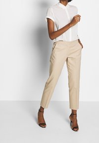 Beżowe dopasowane spodnie z płaskim przodem, bocznymi kieszeniami i wąskim krojem, zestawione z białą koszulą na krótki rękaw z guzikiem. Czarne sandały na obcasie z paskami.