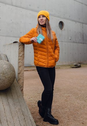 Orange puffer jacket, black jeans, black combat boots. Holding a turquoise wallet with white polka dots, yellow knit beanie. Concrete background.