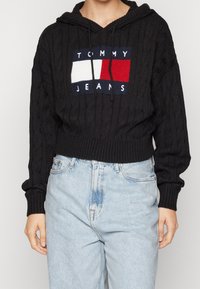 Black cable-knit cropped hoodie with a red, white, and navy "Tommy Jeans" logo, paired with light blue high-waisted jeans.