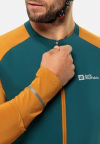 Maillot de cyclisme teal avec épaules orange, fermeture éclair à l'avant, bande réfléchissante sur les manches et logo Jack Wolfskin sur la poitrine.