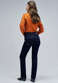 Jeans de un azul oscuro con corte slim y detalles de costura sutiles en los bolsillos traseros, combinados con una blusa naranja de manga larga.