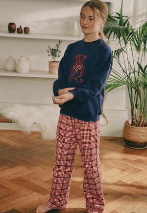 Marineblå sweatshirt med et rødt teddy bear grafisk design, parret med rød- og hvidternet natbukser, lavet af blødt stof. Barfodet på trægulv.