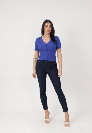 Donna in piedi che indossa una maglietta blu a maniche corte con bottoni e coste, jeans skinny scuri, tacchi bianchi aperti e un tatuaggio visibile sull'avambraccio sinistro.