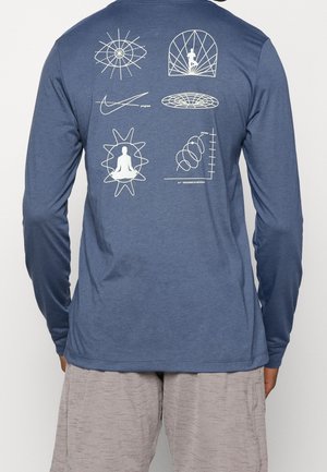 Chemise bleue à manches longues avec dessin en ligne blanche de figures méditant et escaladant, formes abstraites et graphiques avec les mots « progrès » et « ancré dans le mouvement ».