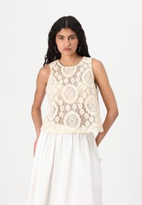 Beige blonde lace crop top med sirkulære utskjæringer og rund hals, båret over en hvit, samlet skjørt, som skaper en lagdelt tekstureffekt.