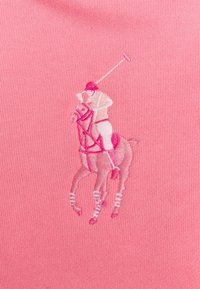 Polo-spelare i hjälm som svingar sin klubba på hästryggen, broderat i nyanser av rosa på en rosa ribbad tygbakgrund.