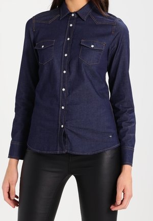 Femme portant une chemise en jean bleu foncé à manches longues avec poches poitrine boutonnées et un pantalon noir ajusté, debout devant un fond uni.
