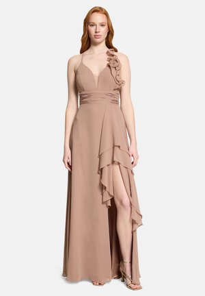 Kobieta ubrana w beżową suknię bez rękawów z kwiatowym zdobieniem na ramieniu, marszczonym stanem i asymetrycznym rozcięciem z falbankami, w połączeniu ze złotymi szpilkami.
