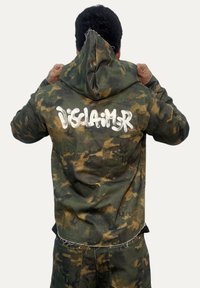 Felpa con cappuccio a motivo mimetico in verde e marrone. Presenta un grande testo bianco "DISCLAIMER" sul retro, con una finitura testurizzata e consumata.