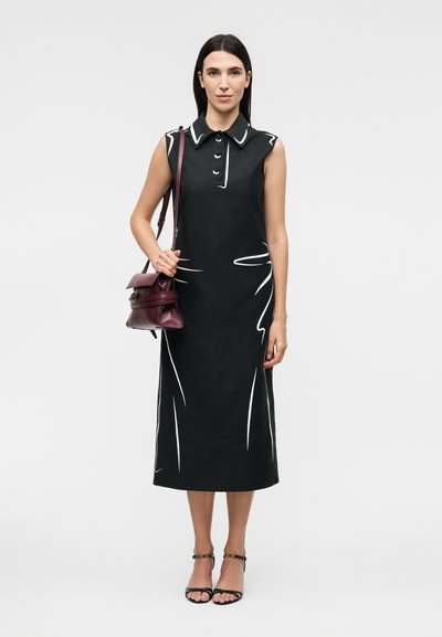 Γυναίκα με μαύρο μίντι αμάνικο φόρεμα με λευκά σχέδια γραμμών, κρατώντας μια μπορντό τσάντα ώμου και φορώντας μαύρες γόβες με λουράκι στον αστράγαλο.