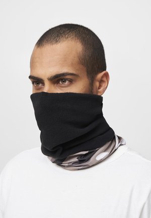 Man with short hair wearing a black fleece neck gaiter over mouth and nose, with a patterned fabric underneath, dressed in a white shirt.