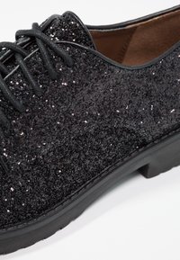 Chaussure noire à paillettes avec lacets lisses et bout arrondi. Tige texturée en matière étincelante, semelle noire épaisse.