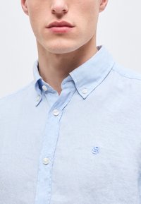 Νεαρός άνδρας που φοράει ένα ανοιχτό γαλάζιο πουκάμισο με μικρό κεντητό λογότυπο στο στήθος, με κουμπιά στον γιακά κλεισμένα.