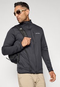 Svart jakke med glidelås og strukturert stoff, med en glidelåslomme og reflekterende logo. Brukt med en ryggsekk og solbriller.