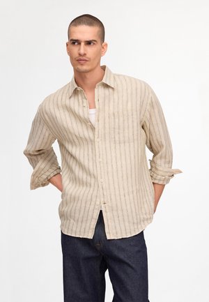 Mand med kort hår iført en beige stribet skjorte med knapper over en hvid undertrøje og mørkeblå jeans, der kigger til siden.