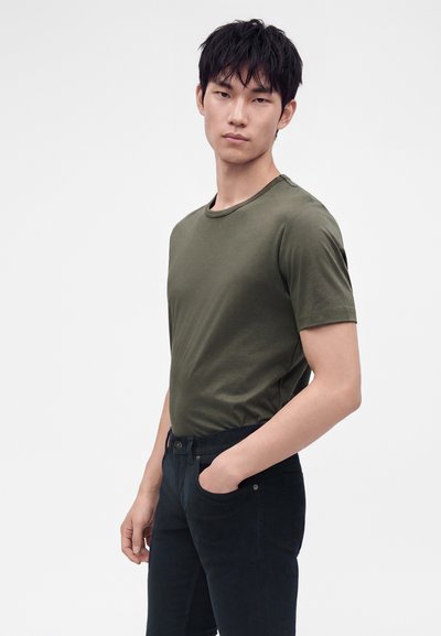 Jeune homme aux cheveux courts noirs portant un t-shirt vert olive et un pantalon noir, debout avec une main dans la poche sur fond blanc.