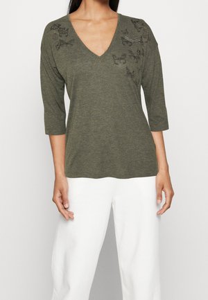 Kobieta w oliwkowozielonym topie z dekoltem w serek z motywem motyli i trzeciowymiarowymi rękawami, w połączeniu z białymi spodniami.