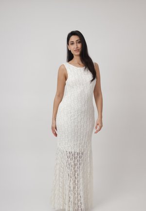 Vrouw met lang donker haar die een mouwloze witte maxi-jurk van kant draagt, staand tegen een effen lichtgrijze achtergrond.