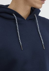 Námornícky modrá mikina vyrobená z mäkkej látky, so šnúrkou so zigzagovým vzorom a kovovými hrotmi. Detailný pohľad na výstrih a kapucňu.