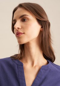 Camicia di lino viola con scollatura rotonda e dettagli di cucitura sottili. La texture liscia e la vestibilità rilassata aumentano il comfort.