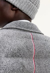 Grau struktuierter Stoffjacke mit einem markanten rot-weißen Akzentstreifen entlang der Naht, ergänzt durch eine hellgraue Strickmütze.