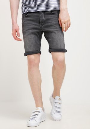Homme portant un short en jean gris retroussé, des baskets blanches à bandes velcro et une chemise rayée, marchant devant un fond uni.