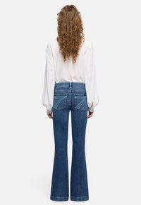 Vrouw met lang, krullend haar, een witte blouse en een high-waist blauwe flared jeans met gestikte achterzakken, van achteren gezien.