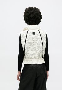 Gesteppte weiße Weste mit wellenförmigem Muster, schwarzen Akzenten, elastischem Saum und hohem Kragen, kombiniert mit schwarzem Langarmshirt und schwarzer Hose.