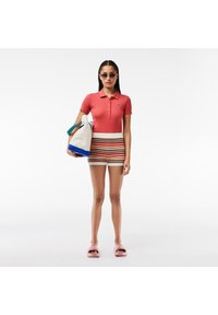 Mujer con una camiseta polo color coral y shorts a rayas, sosteniendo un bolso blanco con acentos en azul y verde azulado, usando gafas de sol redondas y chanclas rosas.
