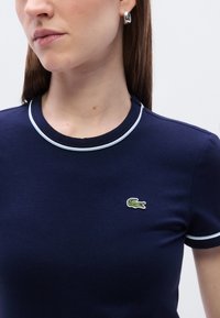 Στενή μπλε μπλούζα σε ν αυτικό μπλε χρώμα με στρογγυλή λαιμόκοψη με λευκή μπορντούρα, μαλακό ύφασμα και μικρό πράσινο και λευκό λογότυπο κροκοδείλου στο στήθος.