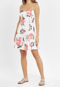 Kvinna i en vit ärmlös klänning med rosa blommönster och silverfärgade platta sandaler, stående mot en helt vit bakgrund.