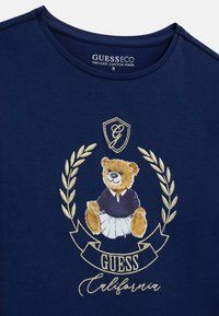 T-shirt in cotone organico blu navy con grafica di un orsetto in un outfit blu circondato da foglie di alloro dorate e testo "GUESS California".
