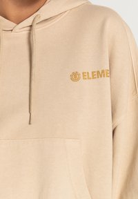 Beige hættetrøje lavet af blødt stof, med en forlommer, snoretræk og gyldent "ELEMENT" logo på venstre brystområde.