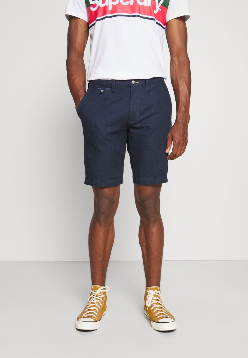 Homme portant un short bleu marine, un t-shirt graphique blanc et des baskets montantes jaune moutarde, debout contre un mur uni.