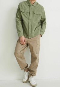 Olivově zelená bunda na knoflíky s dvěma náprsními kapsami, spárovaná s béžovými cargo kalhotami s elastickými manžetami a bílými teniskami s texturovanou podrážkou.