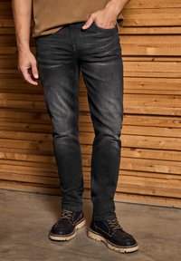 Svarta denimjeans med slim fit, lätt blekning och synliga sömnadsdetaljer, tillsammans med mörka läderstövlar och en brun skjorta, mot en träbakgrund.