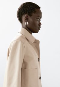 Beige trenchcoat med krave, to knapper og en stor lomme. Model med kort, struktureret hår og en hoop-ørering, vist i profil.