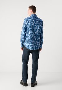 Chemise à boutons bleue à motif floral avec un col évasé, ornée d'un motif de petites fleurs lilas et blanches ; portée avec un jean sombre et des chaussures noires.