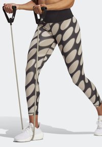 Leggings negras de cintura alta con patrones ovalados en beige, combinados con zapatillas deportivas blancas y bandas de resistencia grises sujetas por los mangos.
