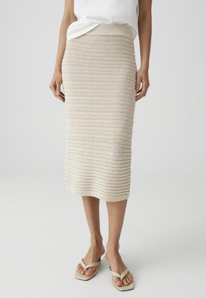 Femme portant une jupe midi beige au crochet associée à un top blanc sans manches et des sandales beiges à talons.