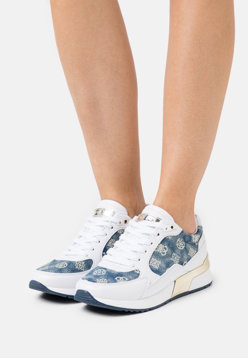 Baskets blanches et bleues à motif avec des lacets blancs portées sur des jambes nues sur un fond clair uni.