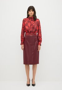 Rood bloempatroon blouse met hoge kraag en ballonmouwen, gecombineerd met een bordeaux leren kokerrok. Zwarte puntenhakken.